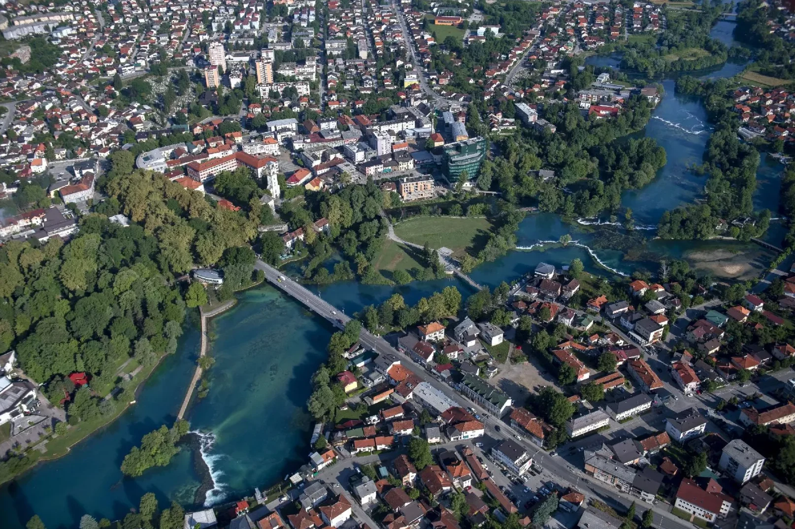Aerial view of a city with a river and waterfalls flowing through it, surrounded by lush green trees and residential buildings.