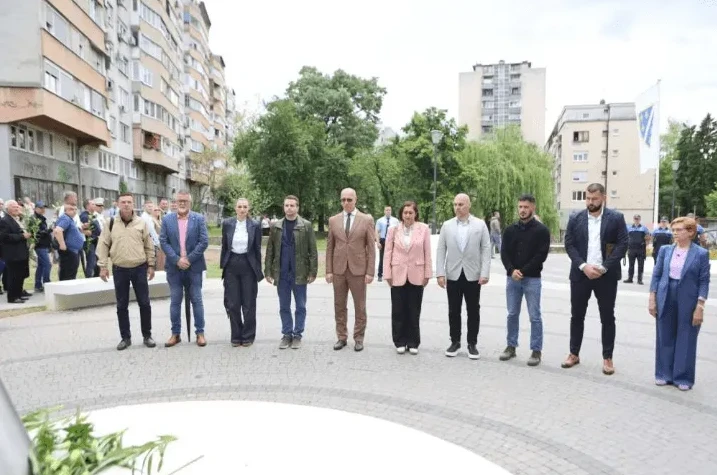 Grupa ljudi, muškaraca i žena u svečanim odijelima, stoji u redu ispred zgrada, u parku, u toku javnog događaja.