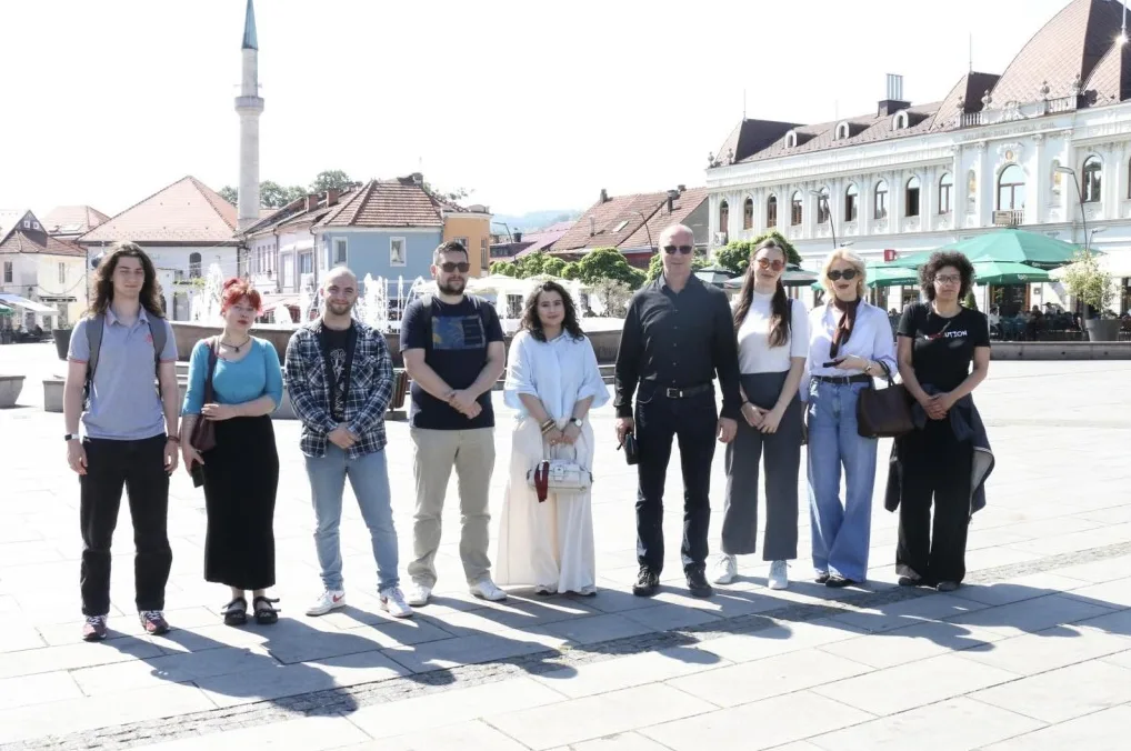 Grupa od devet osoba, pet muškaraca i četiri žene, stoji u redu na popločanom trgu.