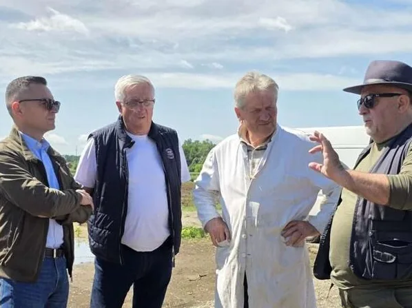 Četiri muškarca razgovaraju vani po sunčanom danu, jedan nosi šešir i gestikulira rukom.