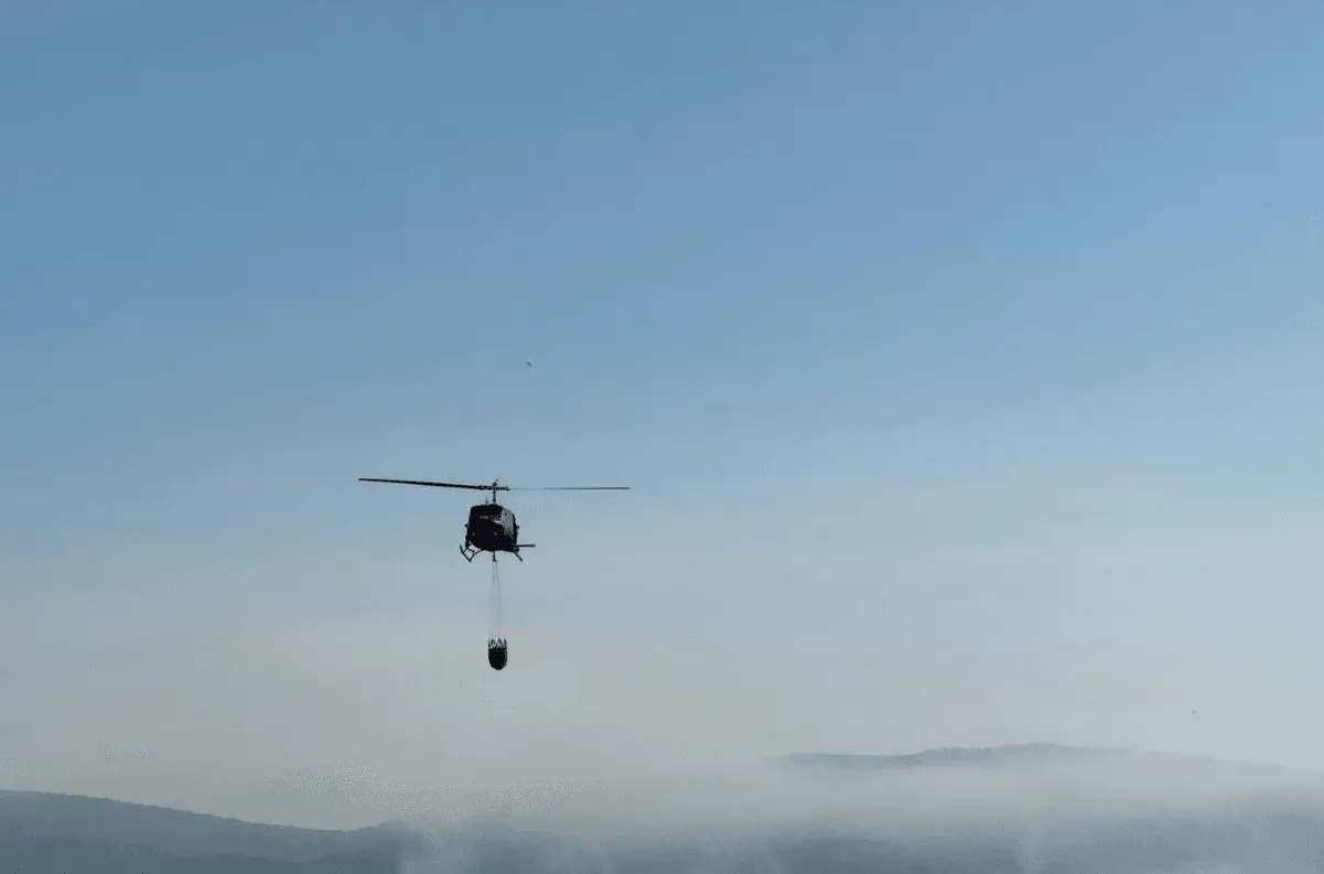 Helikopter nosi veliku kantu napunjenu vodom, prelijećući iznad brdovitog pejzaža prekrivenog maglom.