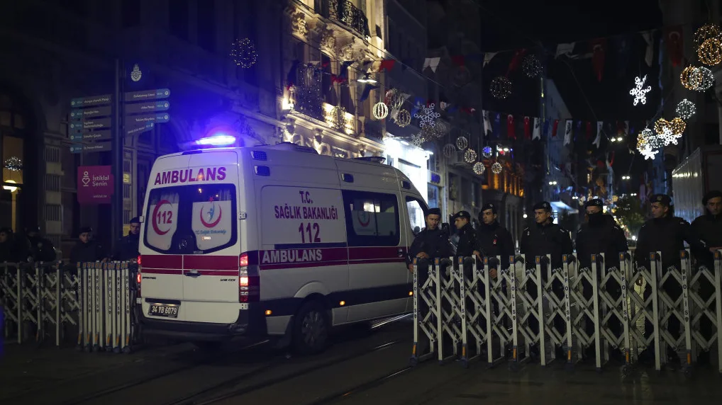 Ambulantna kola sa upaljenim plavim rotacijskim svjetlima stoje iza metalnih barikada na ulici. Pored njih su policajci u crnim uniformama. U pozadini se vide ukrasi i svjetla, te zgrada sa balkonima.