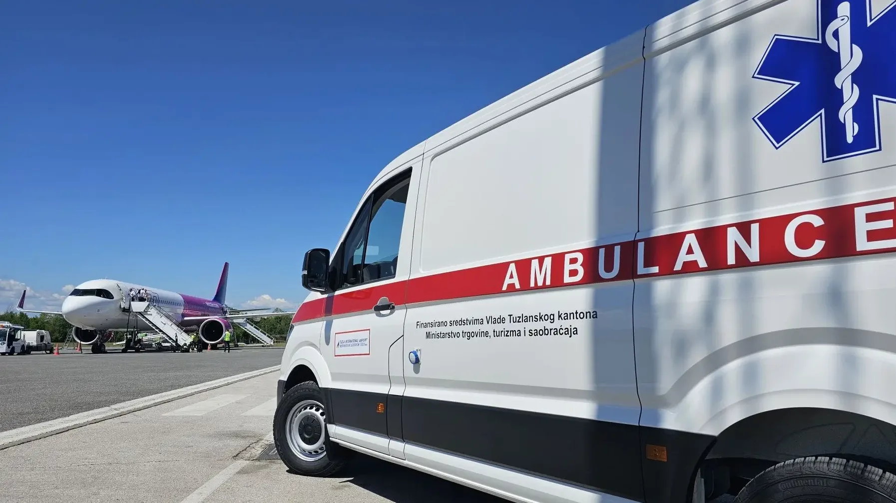 Bijela ambulantna kola sa crvenom trakom i natpisom "AMBULANCE", parkirana na aerodromskom terminalu, sa avionom Wizz Air u pozadini.