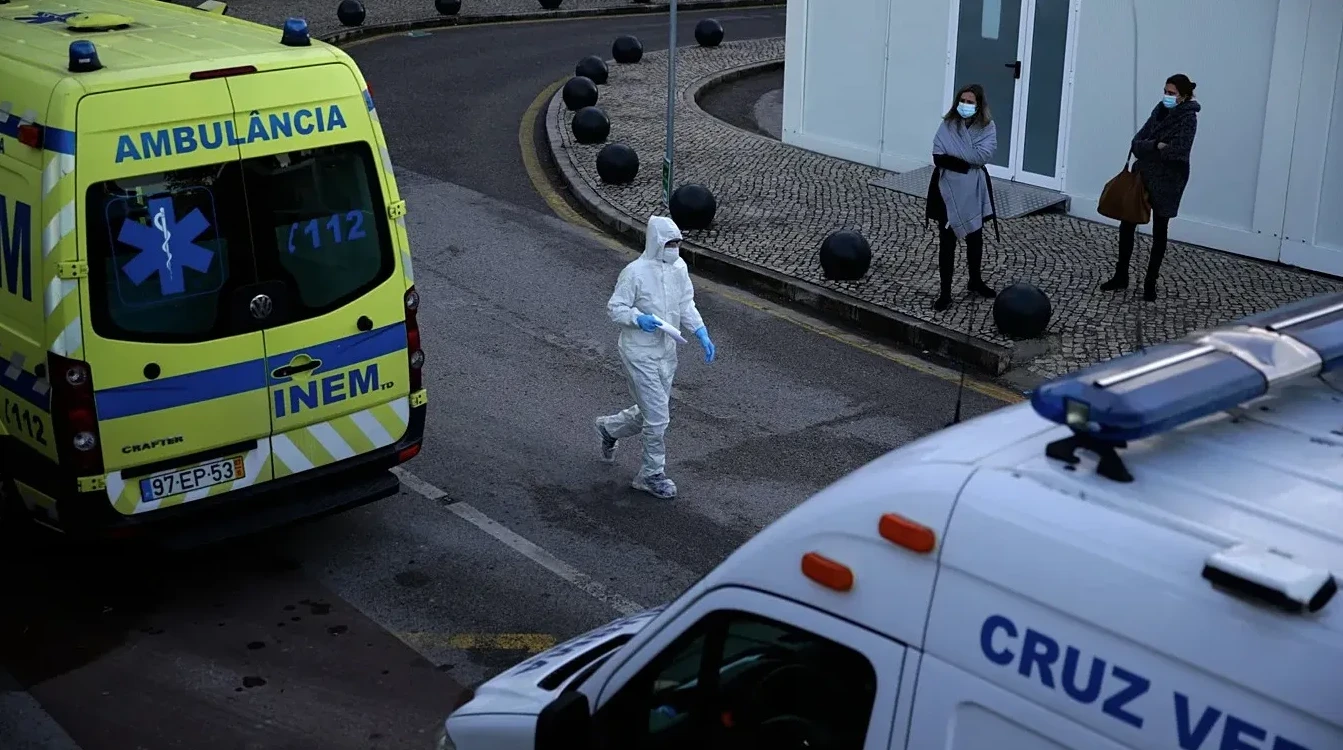 Ambulantna kola hitne pomoći i osoba u potpunom zaštitnom odijelu na otvorenom, s dvije osobe u pozadini koje nose maske.