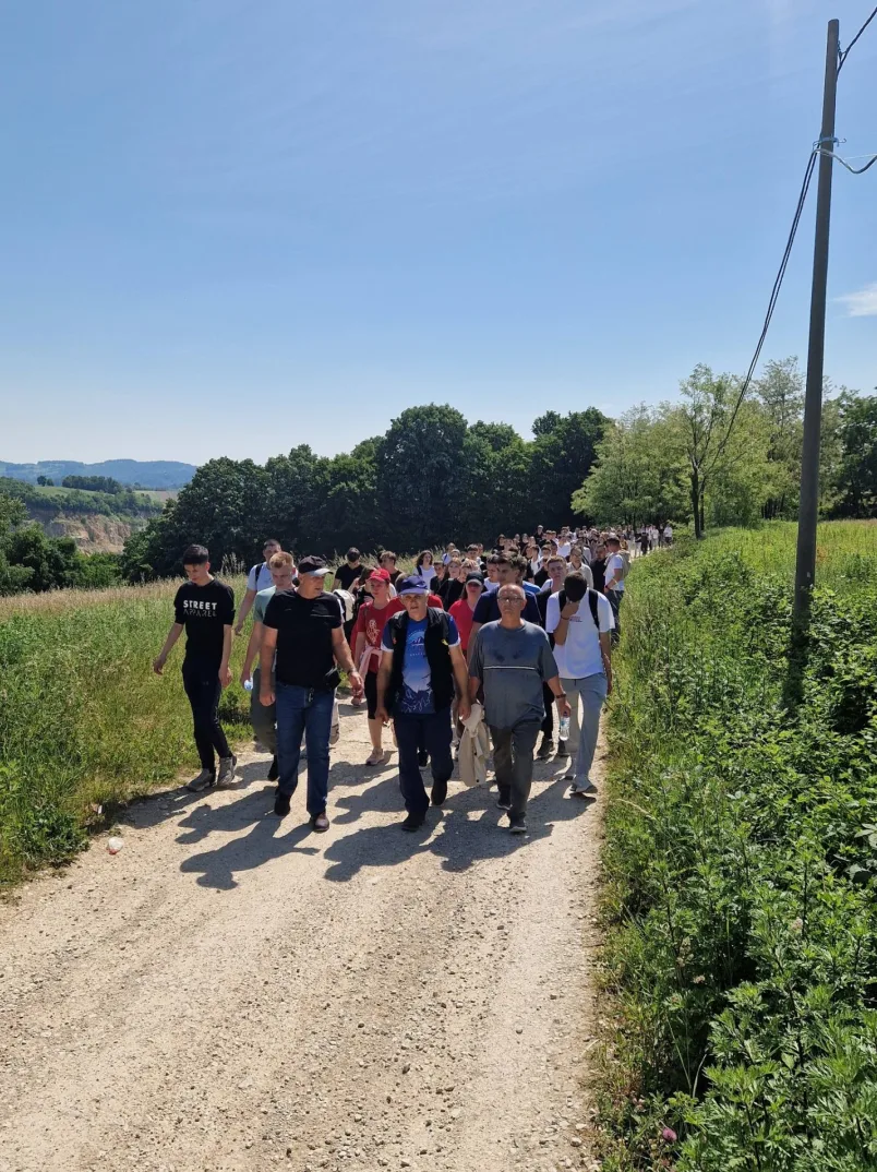 Grupa ljudi pješači makadamskim putem okruženim zelenilom i drvećem.