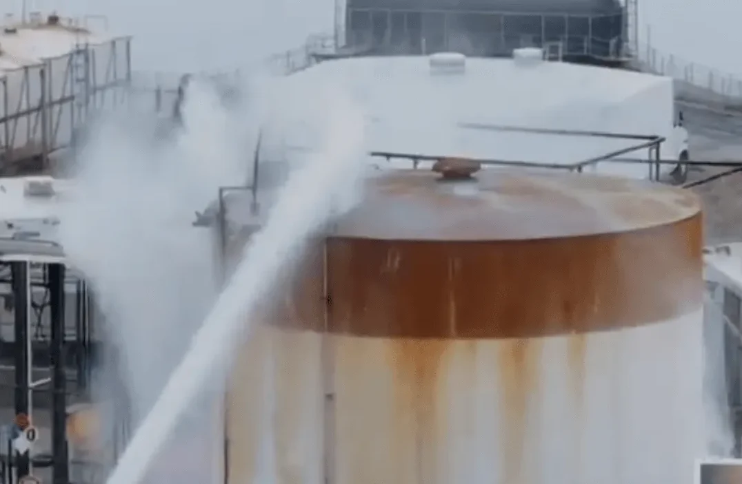 Industrijski rezervoar pod pritiskom ispušta paru ili oblake gasa, sa pratećom infrastrukturom u pozadini.