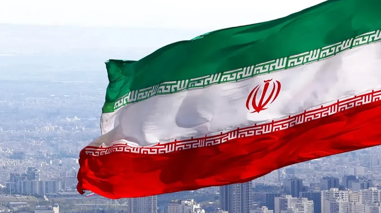 Iranska zastava sa zelenom, bijelom i crvenom prugom, te arapskim pismom i državnim grbom, vijori se na vjetru iznad gradskog pejzaža.