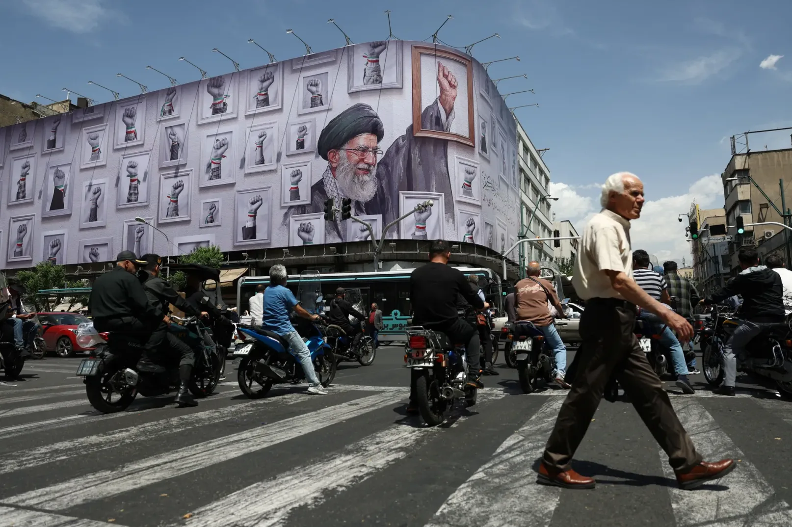 Fotografija prikazuje veliki pano na uglu zgrade u gradu, sa portretom ajatolaha Alija Hamneija okruženim brojnim slikama podignutih pesnica sa iranskim trakama na ručnim zglobovima. Ispod panoa se odvija saobraćaj, sa ljudima na motociklima i automobilima koji prolaze preko pješačkog prelaza.