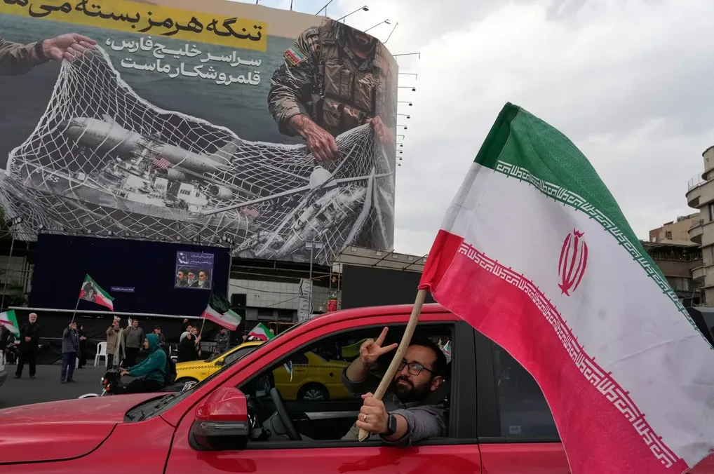 Čovjek u crvenom automobilu maše iranskom zastavom i pokazuje znak mira, dok se u pozadini vidi veliki bilbord sa propagandnom slikom.