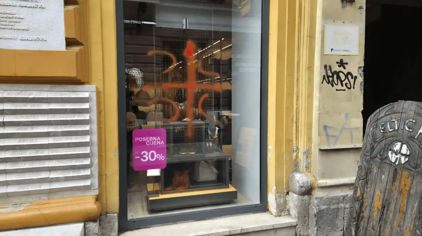 Izlog radnje sa narandžastim grafitom ispisanim sprejem, natpisom "POSEBNA CIJENA do -30%" i drvenom skulpturom sa natpisom "CELTIC AR"