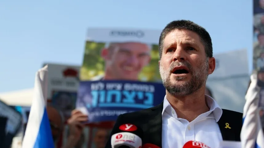 Izraelski političar sa bradom i kratkom kosom govori u mikrofon, noseći crno odijelo preko bijele košulje. Na reveru mu je žuta vrpca. U pozadini se vidi transparent sa slikom mladića i natpisom na hebrejskom, te ljudi koji drže zastave.