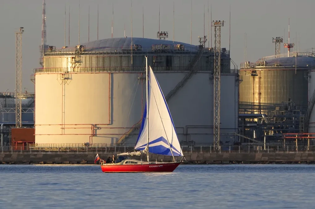 Crveni jedrenjak sa bijelim i plavim jedrima plovi ispred velikih industrijskih rezervoara.