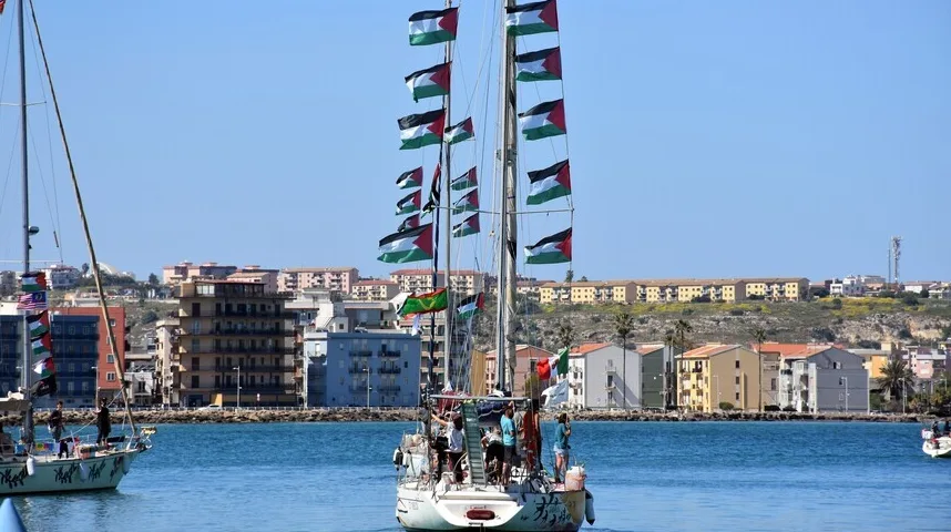 Jedrilica sa zastavama Palestine plovi na moru u blizini obale sa gradskim zgradama u pozadini.