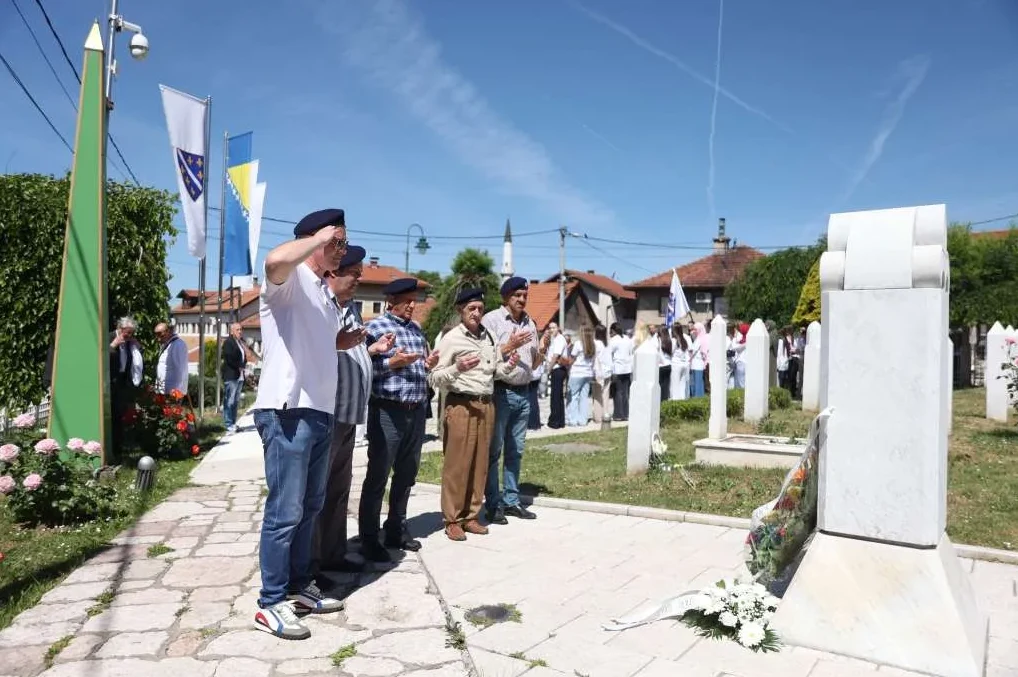Nekoliko muškaraca u beretkama i bijelim majicama na komemoraciji, sa zastavama Bosne i Hercegovine u pozadini.