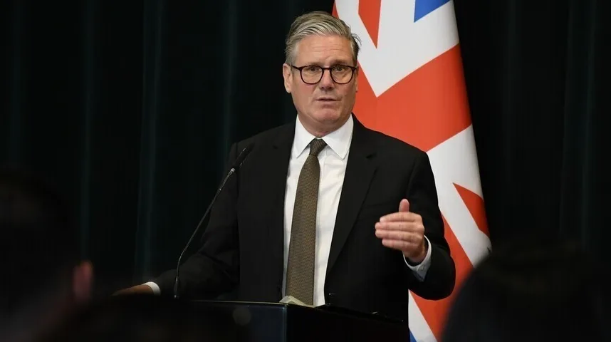 Političar Keir Starmer u crnom odijelu i bijeloj košulji sa smeđom kravatom, sa naočalama, govori ispred zastave Velike Britanije.