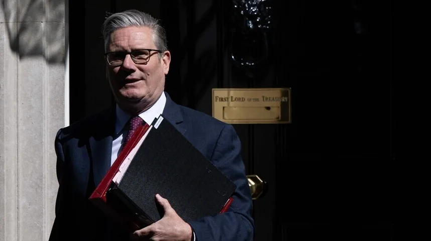 Keir Starmer u tamnom odijelu i crvenoj kravati nosi fascikle ispred zgrade vlade s natpisom 'First Lord of the Treasury'.