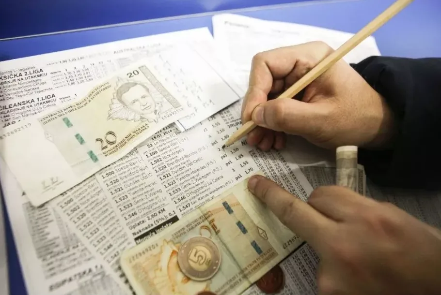 Bliski pogled na ruku koja olovkom obilježava kvote na kladioničarskom listiću, uz novčanice i novčić na stolu.