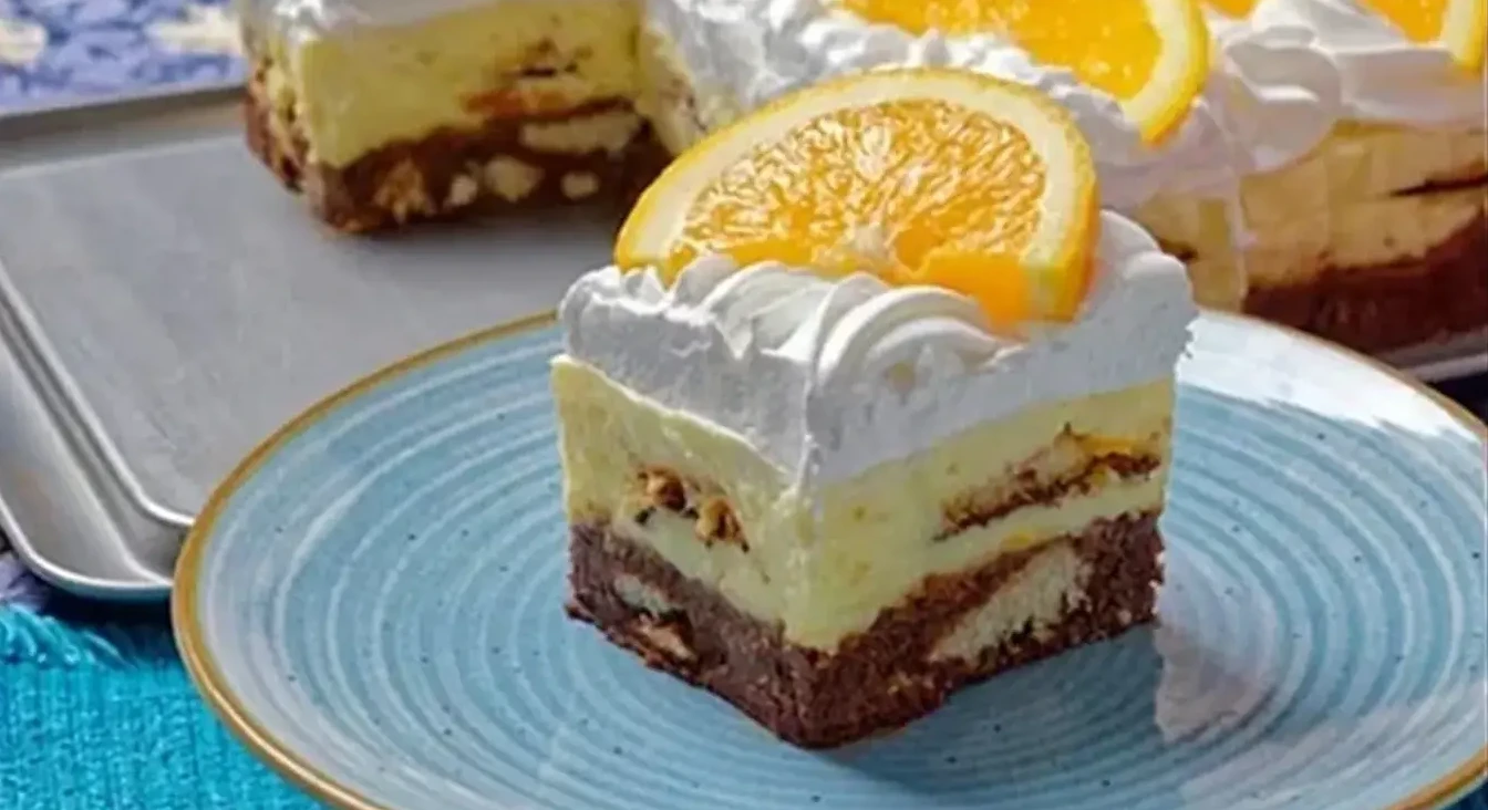 Odrezani komad raskošne torte s više slojeva, ukrašen šlagom i kriškom narandže.