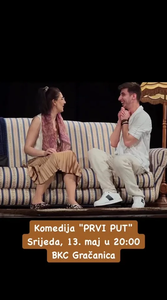 Glumci predstave "PRVI PUT" na sjedećoj garnituri, dama u smeđoj suknji i muškarac u bijeloj košulji, smiju se.