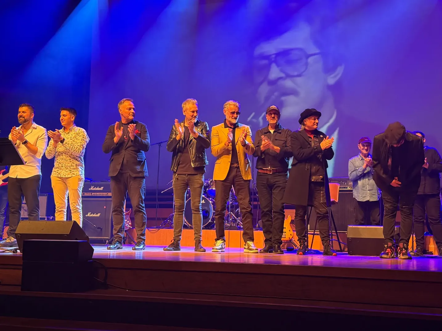 Grupa muzičara aplaudira na pozornici osvijetljenoj plavim reflektorima, dok se na platnu iza njih prikazuje crno-bijela slika starijeg gospodina.