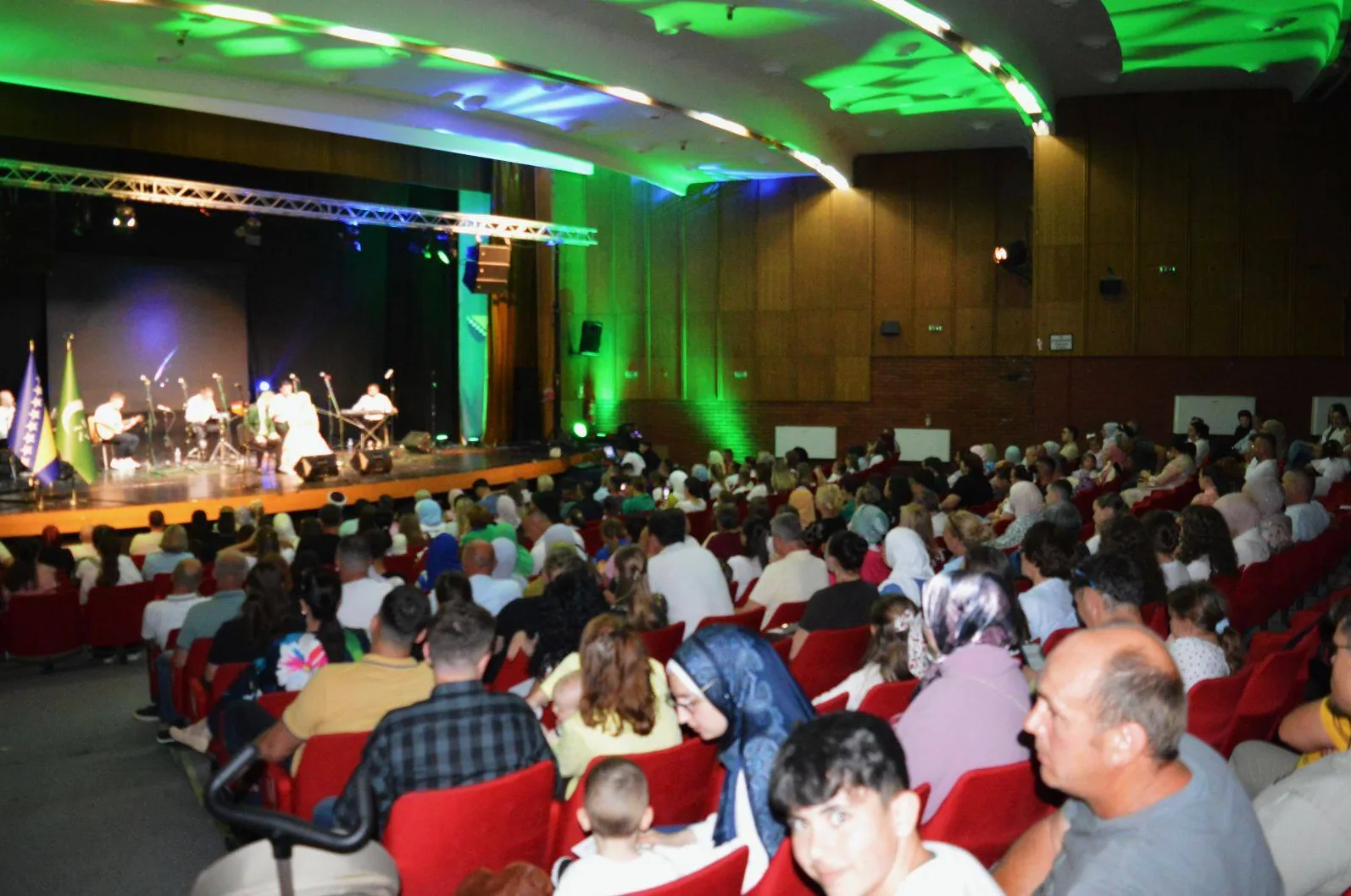 Publika prati koncert u dvorani, na bini muzičari i zastave Bosne i Hercegovine.