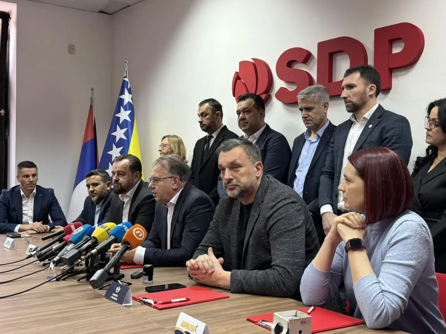 Grupa ljudi sjedi za stolom ispred zastava Bosne i Hercegovine i Srbije, sa logom SDP-a na zidu.
