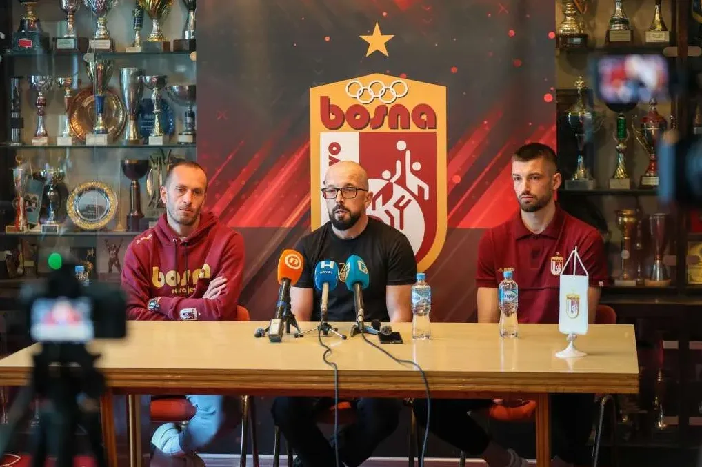 Tri muškarca sjede za stolom ispred panoa sa grbom kluba i peharima u pozadini, na konferenciji za medije.