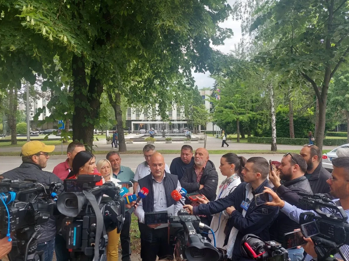 Grupa ljudi, među kojima su političari i novinari, okupila se ispred zgrade na otvorenom, okruženi zelenilom.