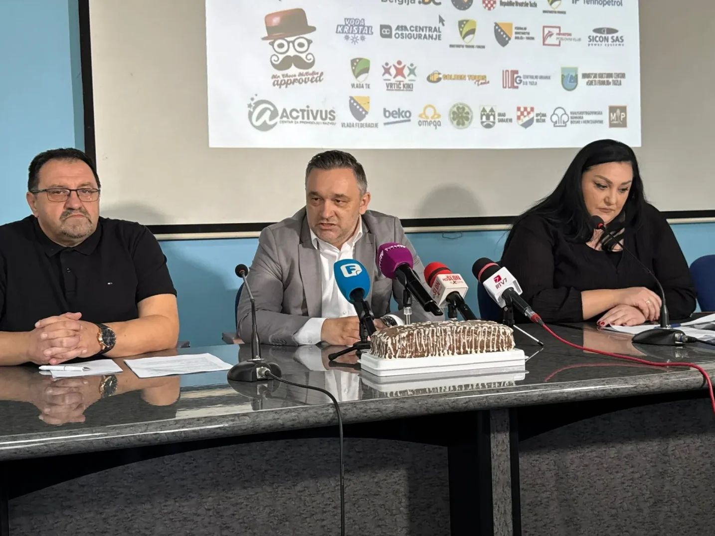 Tri osobe sjede za stolom ispred banera sa logotipima raznih sponzora, dok su ispred njih mikrofoni i torta.