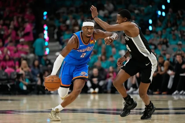 Košarkaš Oklahoma City Thundersa sa brojem 2 na dresu, u plavom šortsu i dresu, dribla loptu dok ga u odbrani čuva igrač San Antonio Spursa sa brojem 4 na crno-bijelom dresu. Utakmica se igra pred publikom u dvorani.