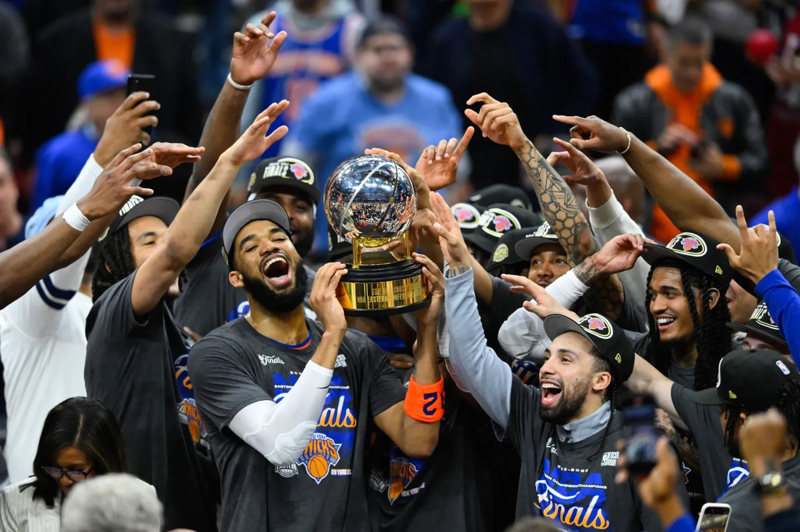 Grupa košarkaša New York Knicksa u majicama "Finals Eastern Conference Champions" slavi podižući veliki trofej u zrak. Igrači nose kačkete i smiju se, a neki od njih drže mobilne telefone i snimaju trenutak.