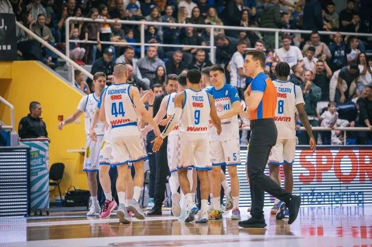 Košarkaški tim u bijelim dresovima sa brojevima i prezimenima na leđima, pozdravlja se sa sudijom na terenu.
