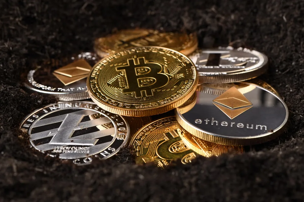 Zlatnici i srebrnjaci bitcoina, ethereuma i litecoina naslagani u tamnoj, hrapavoj podlozi, vjerovatno zemlji ili tlu.