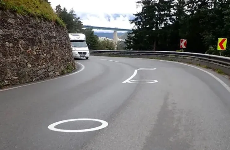 Bijeli autodom vozi krivudavim planinskim putem sa kružnim bijelim oznakama na kolovozu.