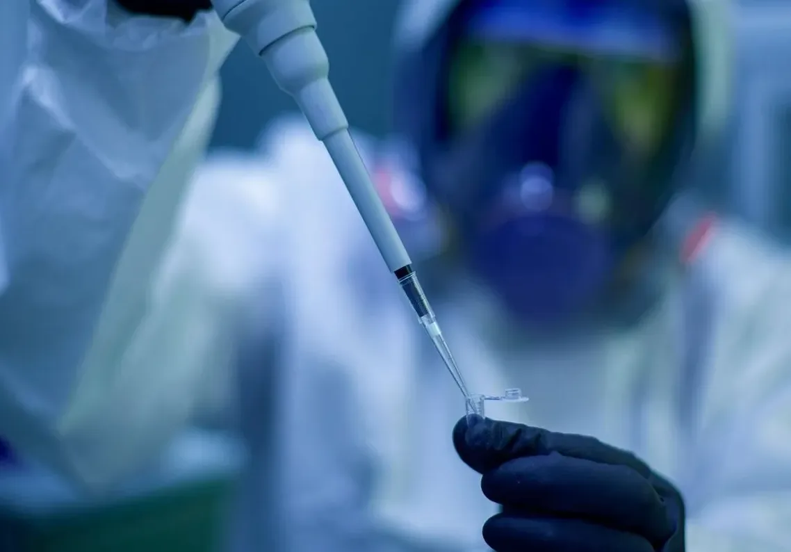 Laboratorijski radnik u zaštitnom skf odijelu i gas maski precizno koristi pipetu za dodavanje tečnosti u malu epruvetu.