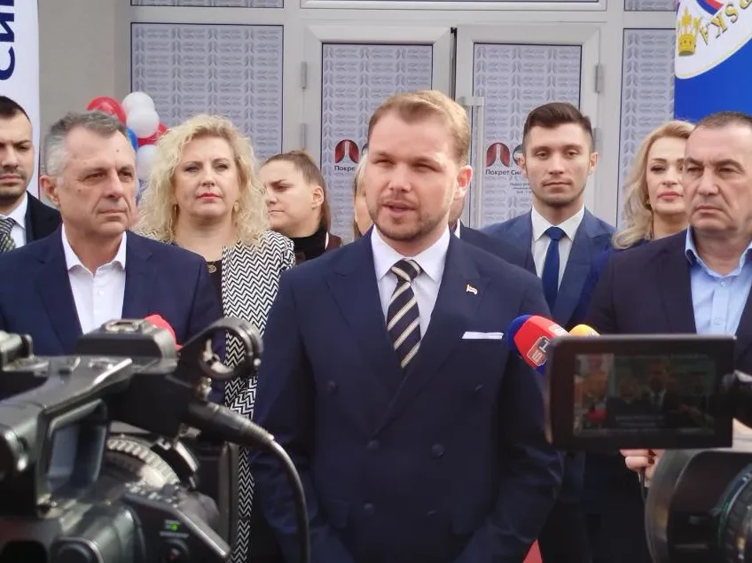Grupa zvaničnika prisustvuje otvaranju, fokus na muškarcu u tamnoplavom odijelu koji govori u mikrofon, okružen medijskom ekipom