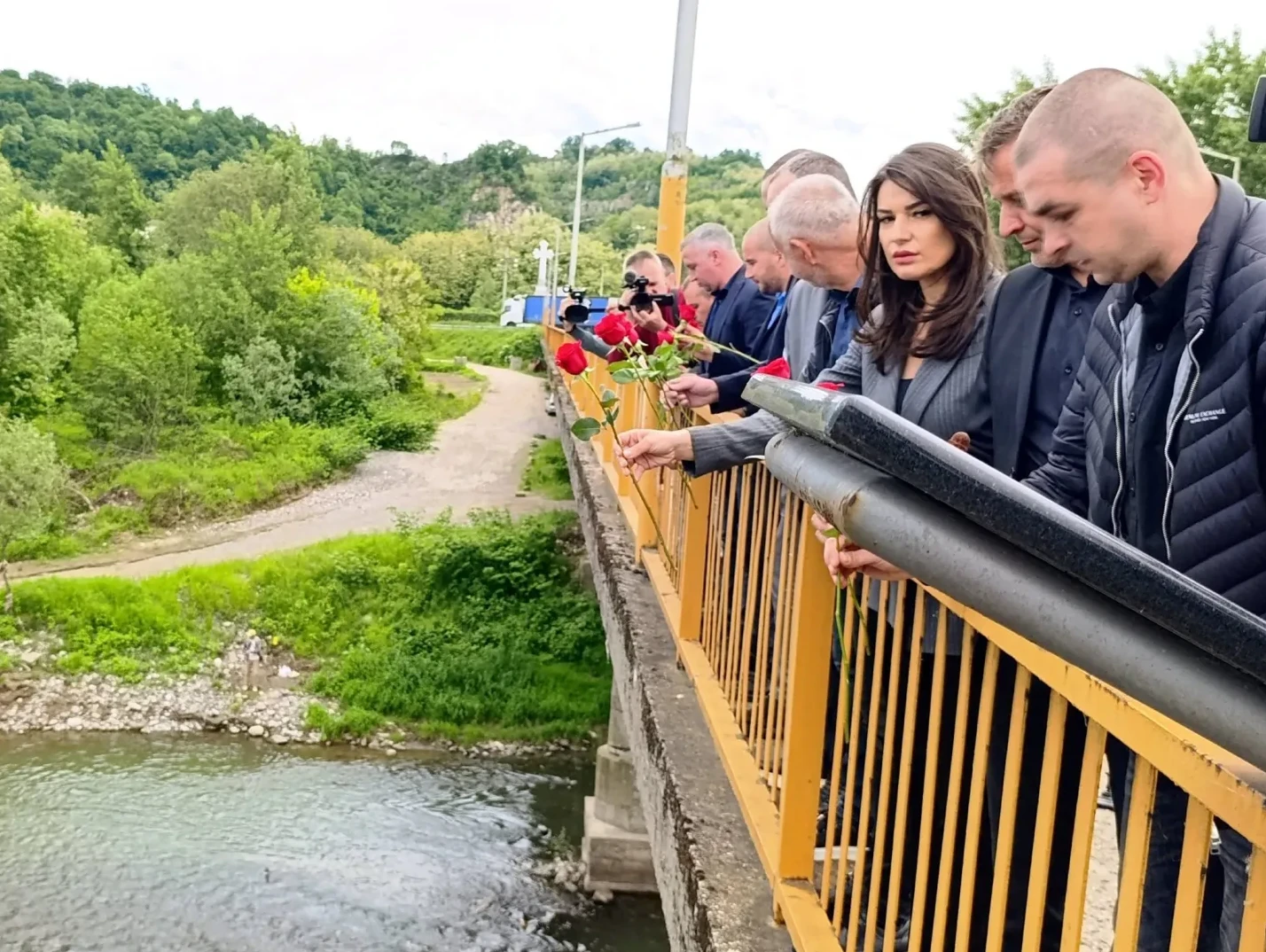 Grupa ljudi, među kojima i jedna žena u sivom odijelu, stoji na žutom mostu i spušta crvene ruže preko ograde u rijeku. U pozadini se vidi zelenilo i brdovit krajolik.