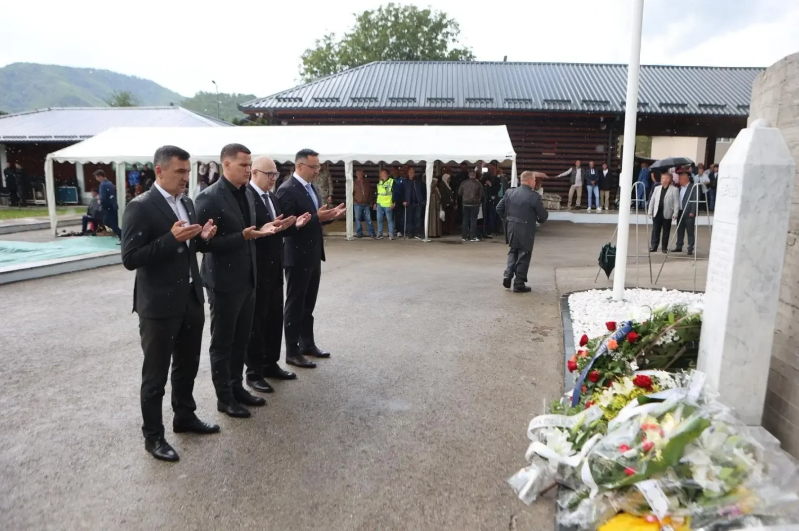 Grupa muškaraca u crnim odijelima drži ruke u molitvi ispred spomen-obilježja prekrivenog cvijećem i vijencima. U pozadini su vidljive šatorske konstrukcije i okupljeni ljudi.