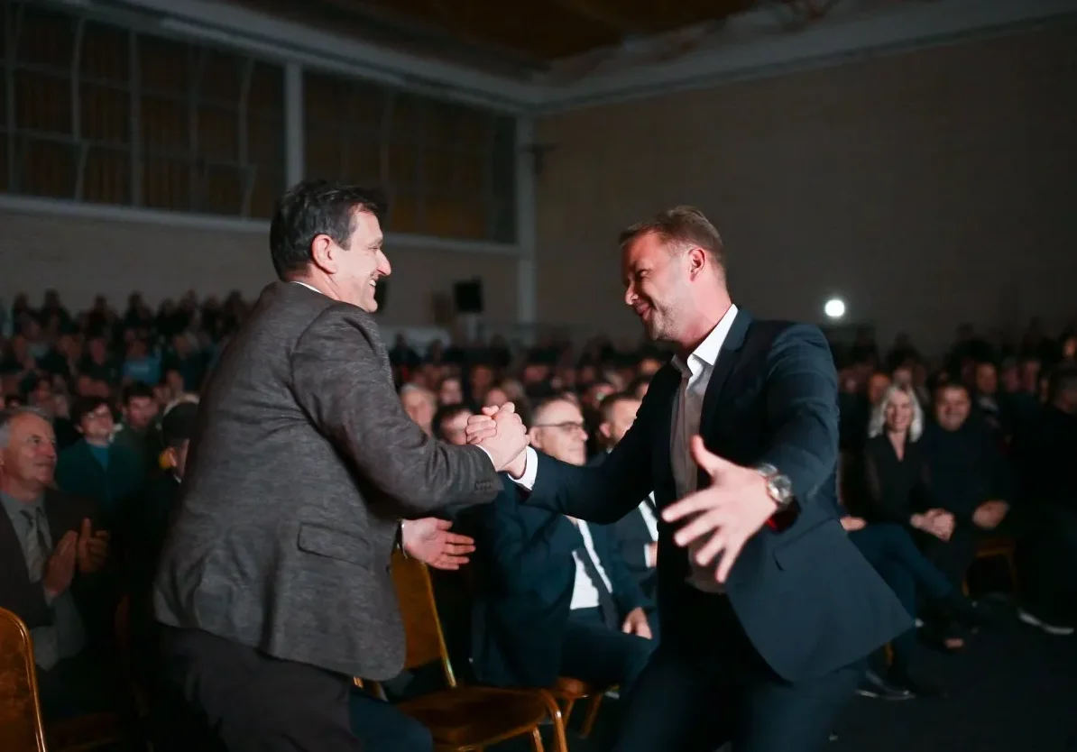Dvojica muškaraca u odijelima se rukuju na pozornici pred publikom.