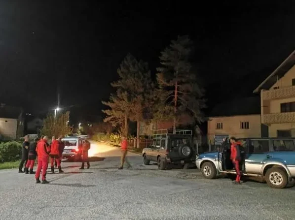 Grupa ljudi u crvenim vatrogasnim odijelima okupljena je pored bijelog terenskog vozila na asfaltiranoj podlozi, pod okriljem noći. U pozadini se naziru drveća i osvijetljena kuća, te drugi parkirani terenci.