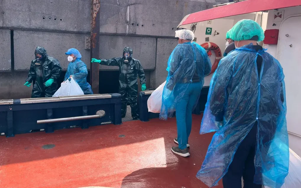 Tim u punoj zaštitnoj opremi, uključujući odijela, maske i rukavice, na palubi.