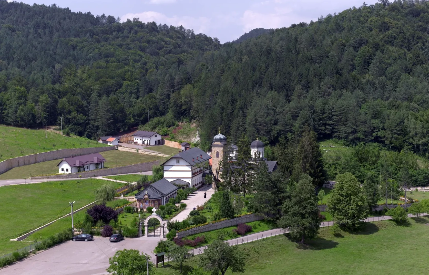 Prizor manastira i okolnih objekata okružen bujnom zelenom šumom i zelenim poljima.