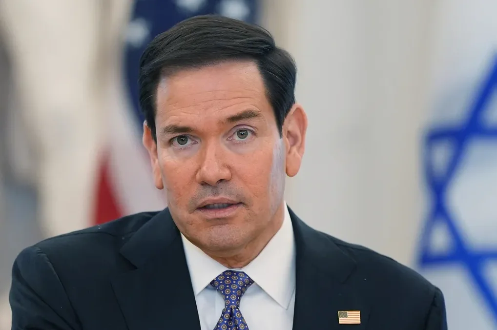 Američki senator Marco Rubio u tamnom odijelu, bijeloj košulji i plavoj kravati sa uzorkom, sa zastavom Sjedinjenih Američkih Država i Izraela u pozadini.