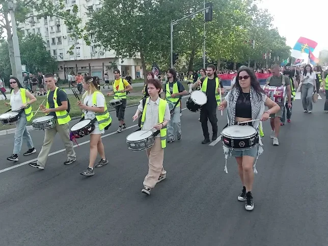 Grupa mladih ljudi u reflexivnim prslucima maršira ulicom i svira na bubnjevima, dok se u pozadini vide zastave Srbije.