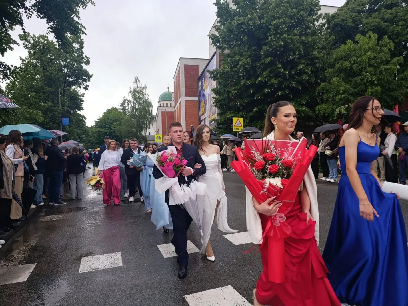 Maturanti sa cvijećem prolaze ulicom, obučeni u svečane haljine i odijela, dok ih neki prate sa kišobranima.