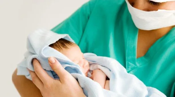 Medicinska sestra u zelenoj uniformi i sa maskom na licu nježno drži tek rođenu bebu umotanu u plavi peškir.