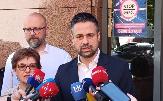 Premijer Federacije BiH, Nermin Nikšić, obraća se medijima na konferenciji za novinare, držeći mikrofon. Pored njega su ministrica finansija Federacije BiH, Jelka Milićević, i premijerov savjetnik. U pozadini se vidi plakat sa natpisom "STOP ŠVERCU" i brojem telefona.