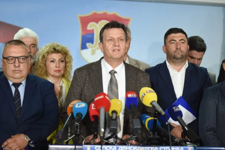 Grupa političara, među kojima prednjači muškarac u tamnosmeđem sakou koji se obraća okupljenima, stoji iza govornice s brojnim mikrofonima. U pozadini se vidi grb.