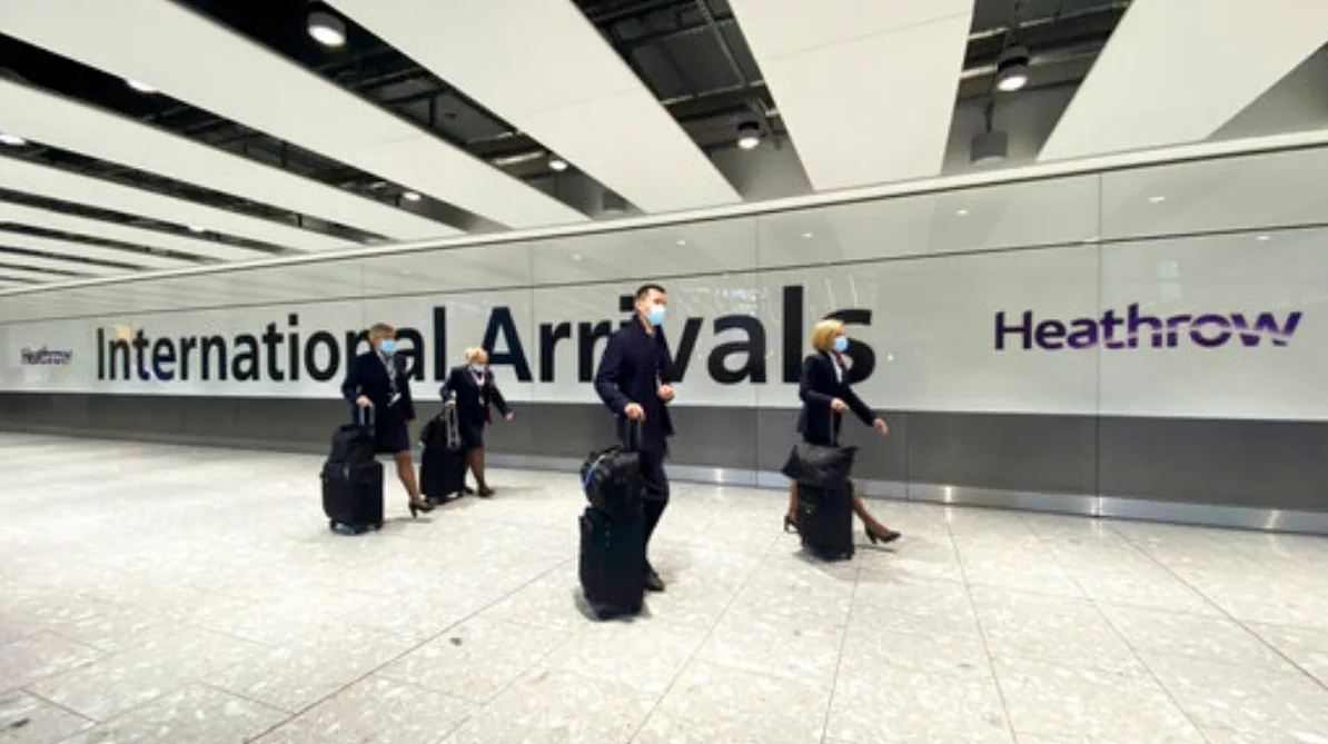 Grupa putnika sa prtljagom, svi nose zaštitne maske, prolazi kroz međunarodni dolazni terminal aerodroma Heathrow.