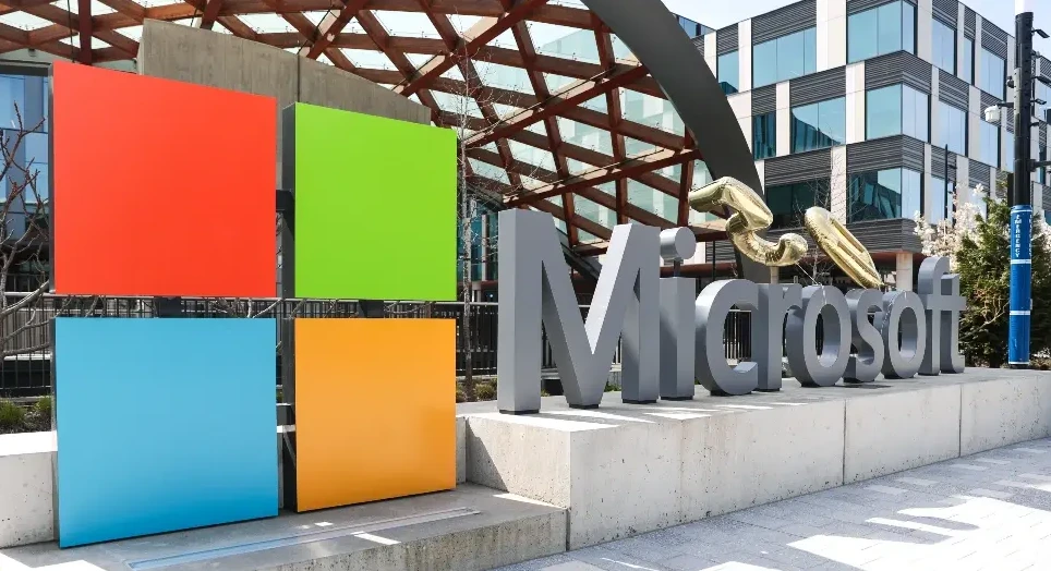 Slovo "Microsoft" ispisano velikim sivim slovima, sa živopisnim logoom kompanije od četiri kvadrata - crvenim, zelenim, plavim i narandžastim, ispred moderne zgrade.